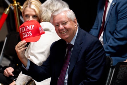 An older man grins while holding a Trump cap.