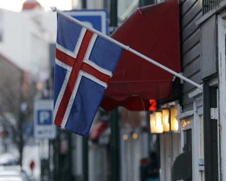 Icelandic flag with its asymmetric cross hangs diagonally outside a wooden shop with four vertical lights behind it