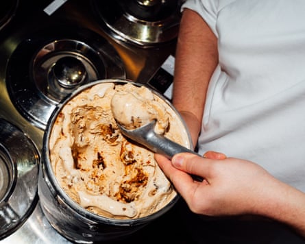 Someone in a white T-shirt scoops a ball of brown and cream ice-cream from a large metal container
