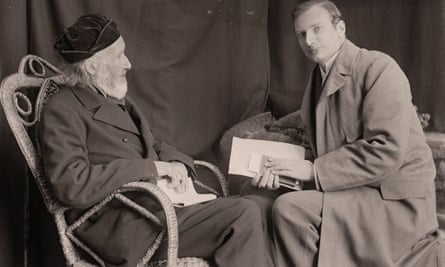 Two men sit opposite one another in chairs with their knees touching. Both have books in their hands and are wearing smart clothing.