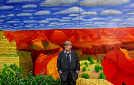David Hockney in glasses and a cap stands in front of his vivid painting of red canyon landscapes