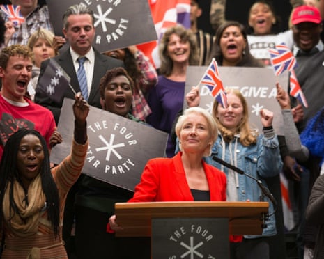 Emma Thompson stands at a podium before a crowd waving Union Jack flags and holding signs