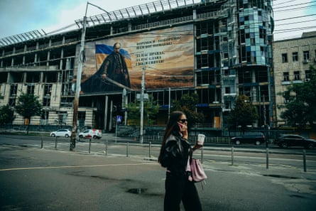 Young woman in sunglasses walks down deserted road, with a bombed out building covered with advertising behind her