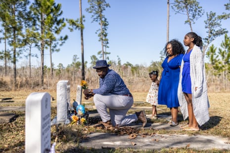 A man places flowers on a tombstone, his two daughters and wife stand behind him