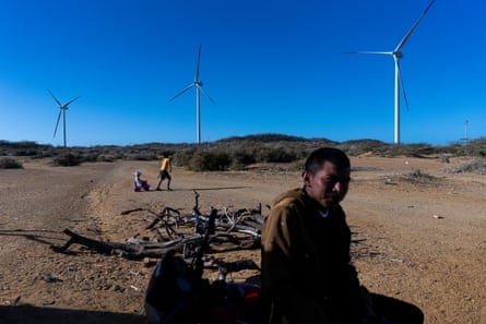 A man looks at the camera while children play. Wind turbines loom behind him