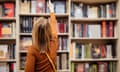 A young girl reaches for a book between bookshelves in a library.