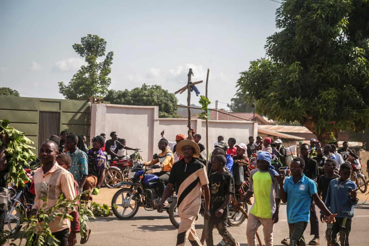 A crowd of people walk or ride motorcycles along a road, two of them holding up makeshift wooden crosses