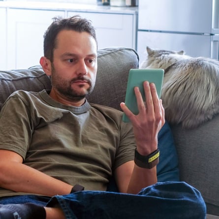 A man on a sofa reads a Kindle e-reader, with a fluffy cat beside him
