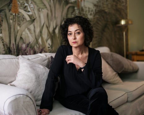 She sits on a cream sofa, dressed in black, looking at the camera, her right arm leaning on the sofa cushion, left left arm held in front of her torso