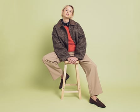 Woman sits on stool wearing dark brown jacket and orange top.