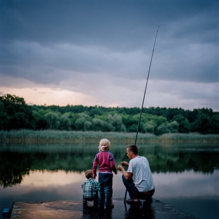A man with a fishing rod crouches down next to a young girl and toddler boy at the side of a lake