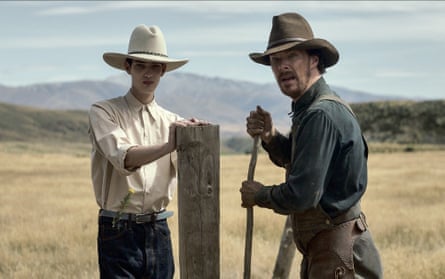 Kodi Smit-McPhee and Benedict Cumberbatch standing by a post wearing cowboy outfits in The Power of the Dog