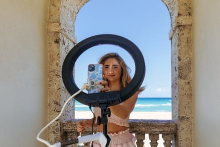 A girl sets up her smartphone on a ring light in front of an archway that looks out onto a bright blue ocean scene.