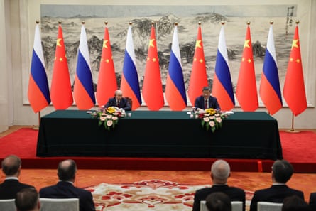 Vladimir Putin and Xi Jinping sitting at a long table with flags behind them