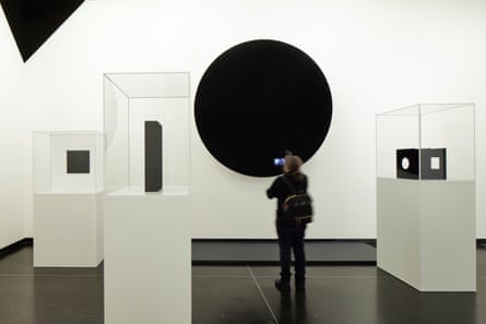 A gallery visitor take a photo on his mobile phone of a large black disc in a room containing other black sculptures