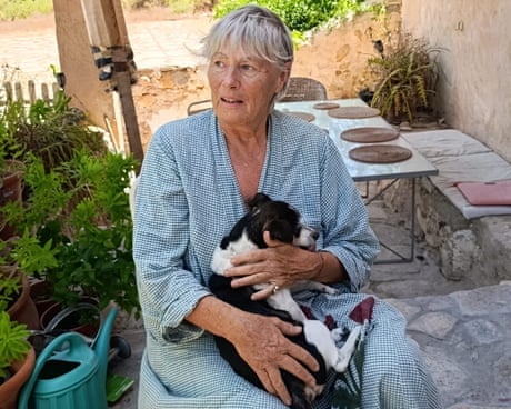 Jeanne Henny holds a black and white dog on her lap while sitting on a stone patio with plants and a table behind her