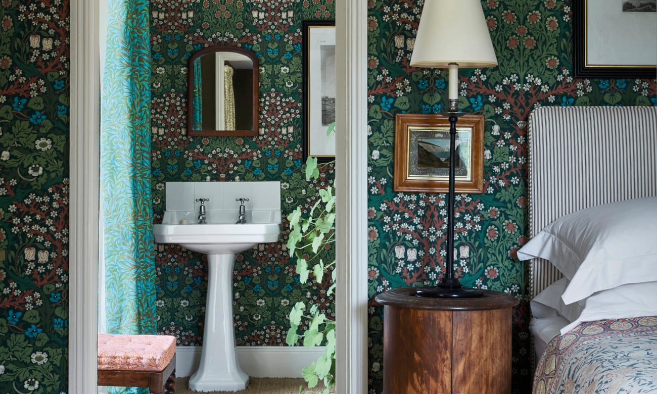 A bedroom with green patterned wallpaper, and a full-length mirror reflecting it
