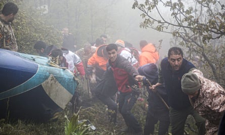 Search and rescue teams work around the wreckage of the helicopter: men are seen forming a chain up the steep hillside, holding a rope; they are wearing anoraks and some are in hi-vis jackets. A corner of the helicopter can be seen on the ground. There is fog in the background and visibility in the area is poor.