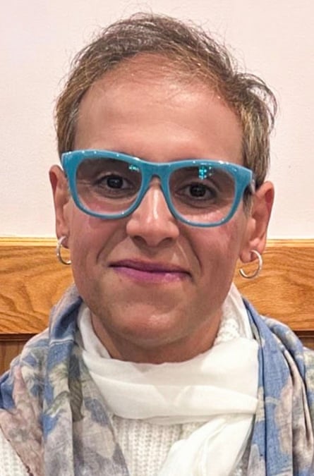 Middle-aged white/Middle-Eastern person with short bright, hoop earrings, and blue-framed glasses