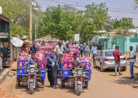 Several pick-up motor-tricycles carry bags of what appear to be food along a sandy dirt road in an African town