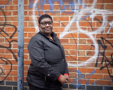 Ros Griffiths stands against a brick wall with graffiti on it. She has close-cropped hair and wears black glasses and a black anorak.