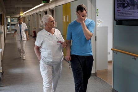 A nurse with short hair in a ponytail and a short-sleeved uniform walks down a hospital corridor with a doctor in a T-shirt holding his glasses in his right hand and putting his left hand over his eyes