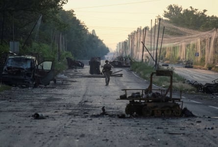 Soldier walking along road amid vehicle debris, with netting overhead.