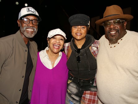 Against a dark background, the four of them stand together and pose for the camera, smiling