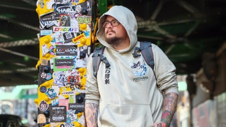 A man with his hood up leans against a post covered in stickers.