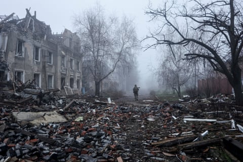 A soldier, in the centre of the picture, walks away from the photographer down a rubble-strewn street with a destroyed house to the left