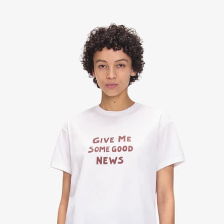 A model wearrs a white short-sleeved T-shirt with the words “GIVE ME SOME GOOD NEWS” in rust coloured lettering.
