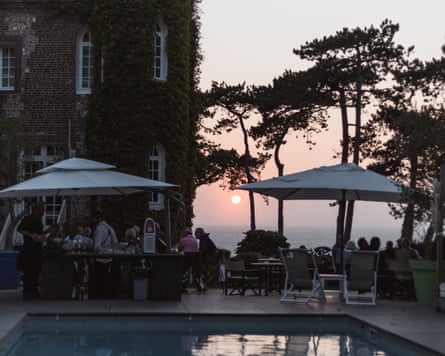 A swimming pool at dusk with people sitting around tables under umbrellas and pine trees, next to an ivy-covered building with a turret.