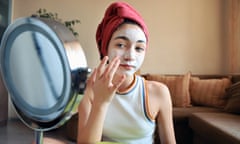 A teenage girl with a towel wrapped around her head, wearing a white vest, applies a white face mask, while looking in a mirror.