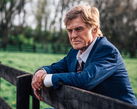 Robert Redford in a blue suit and white shirt leans over a wooden fence in a field with trees in the background.