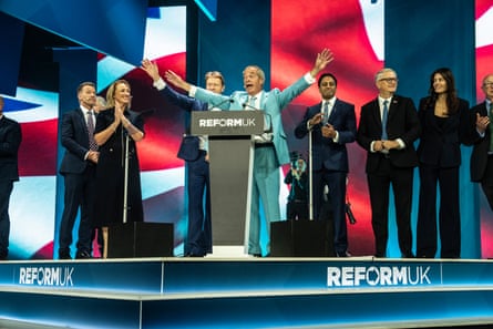 Nigel Farage standing at the podium in his pastel turquoise suit with his arms spread in front of other Reform representatives