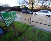 Two cars stuck in flood water, as a sign in the foreground reads 'Please drive carefully – ducks crossing'