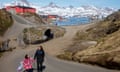 A woman and child hold hands as they walk on a street in the town of Tasiilaq, Greenland, with red buildings, the sea and snowy mountains in the background