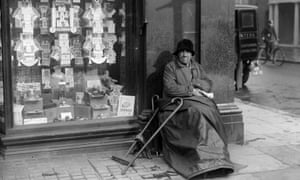 An elderly woman sits by a shop window full of adverts for cigarettes, 1935.
