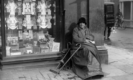 An elderly woman sits by a shop window full of adverts for cigarettes, 1935.