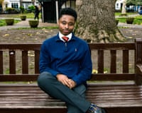 A young man wearing a bright blue quarter-zip jumper over a white shirt and striped tie, with suit trousers, sitting on a bench in a muddy park with a tree behind him