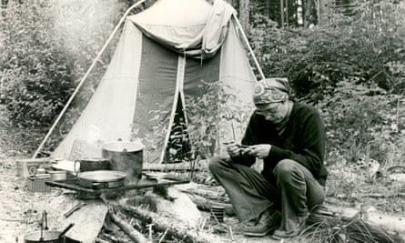 Annie Proulx, hair in a kerchief, sitting on a log in front of a tent and looking down