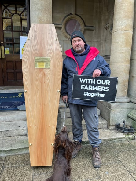 Christopher Marchment stands next to the upright coffin. He is holding a sign that reads 'with our farmers #together' and his brown working cocker spaniel Grouse is sitting facing him