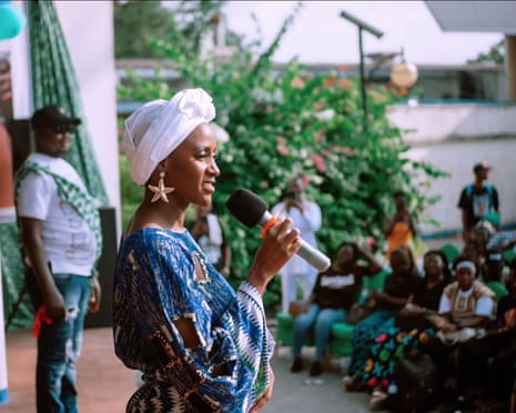 A young African woman in a headdress and loose gown speaks into a mic at an outdoors rally