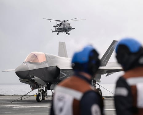 Helicopter comes into land, with fighter jet on flight deck and two service personnel in foreground