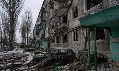 A destroyed car is seen in front of a heavily bombed residential building