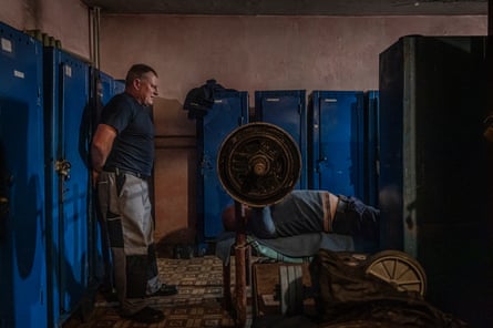 One firefighter leans against a locker while a colleague lifts a large set of weights in front of him