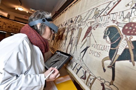 A woman in white coat and protective visor, holding a tablet, inspects the tapestry closely; a detail showing a soldier on horseback and a footsoldier with weapon is seen.