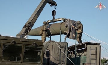 Two soldiers standing on top of a military vehicle position a missile being hoisted into place by a crane