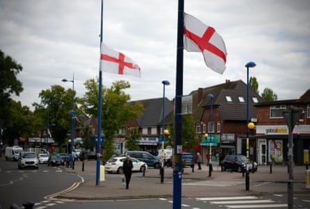 Red and white England flags fly from lamp-posts in a suburban street with shops and a pedestrian road crossing in the background.