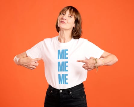 A photo of Zoe Williams in front of a bright orange backdrop. She is wearing a white graphic T-shirt with the words ‘ME ME ME’ in bold blue font, and is pointing to the words.